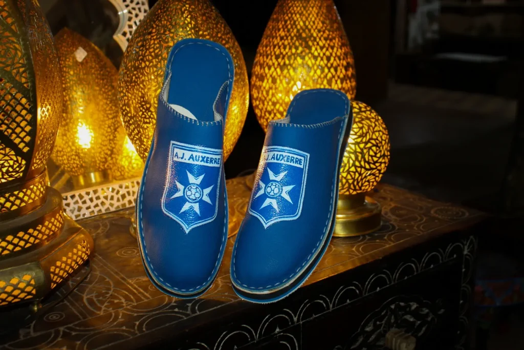 Babouche AJ Auxerre en cuir bleu avec logo peint à la main, posée sur une table orientale artisanale décorée de lampes marocaines, symbole d’élégance et de fabrication traditionnelle.