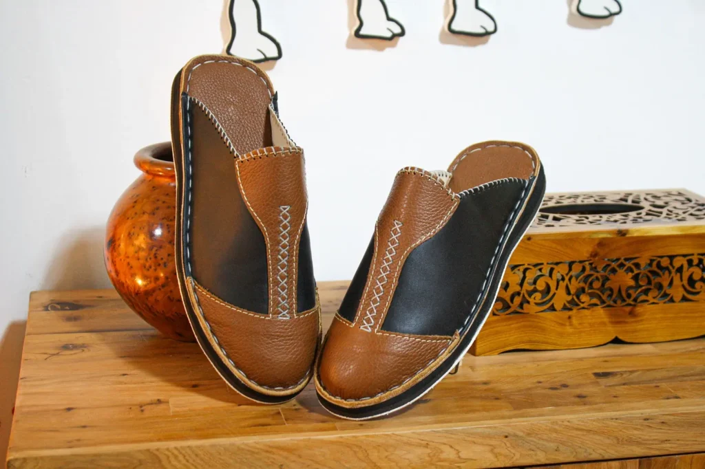 Babouche en cuir pour homme marron et noir faite main, posée sur table en bois avec décor artisanal marocain
