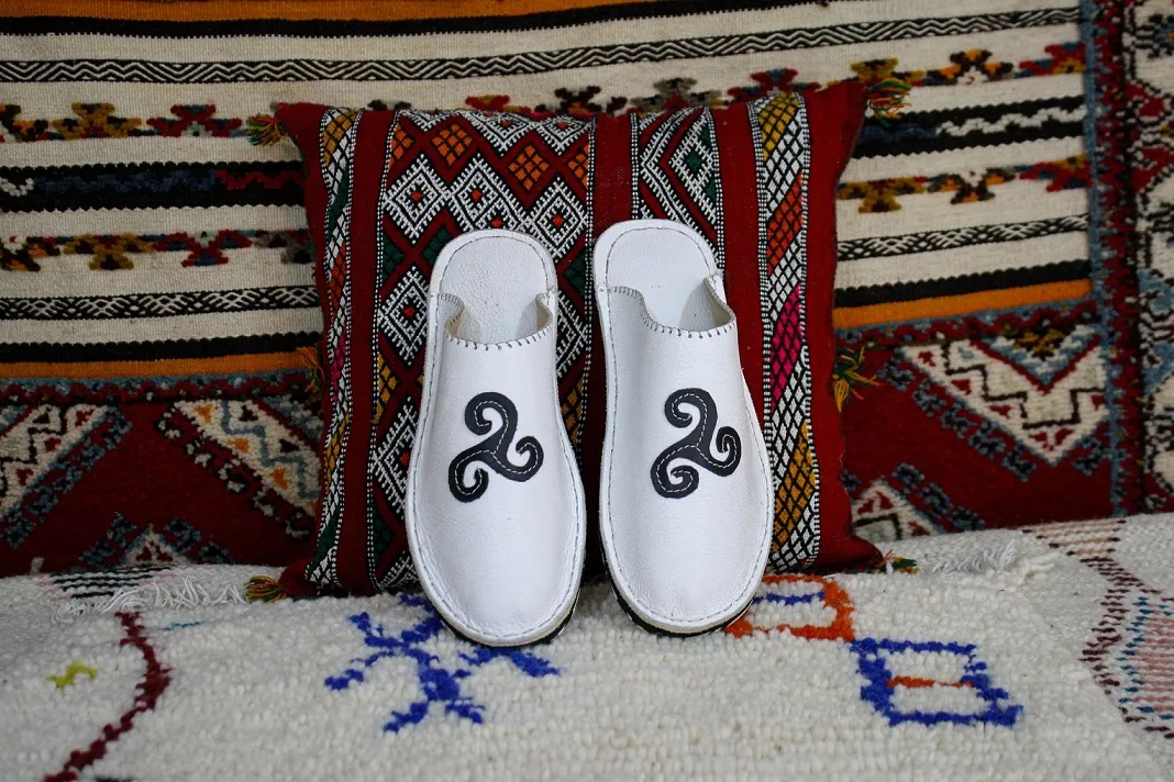 Babouche Bretagne blanche en cuir avec motif breton noir, posée sur un coussin berbère coloré